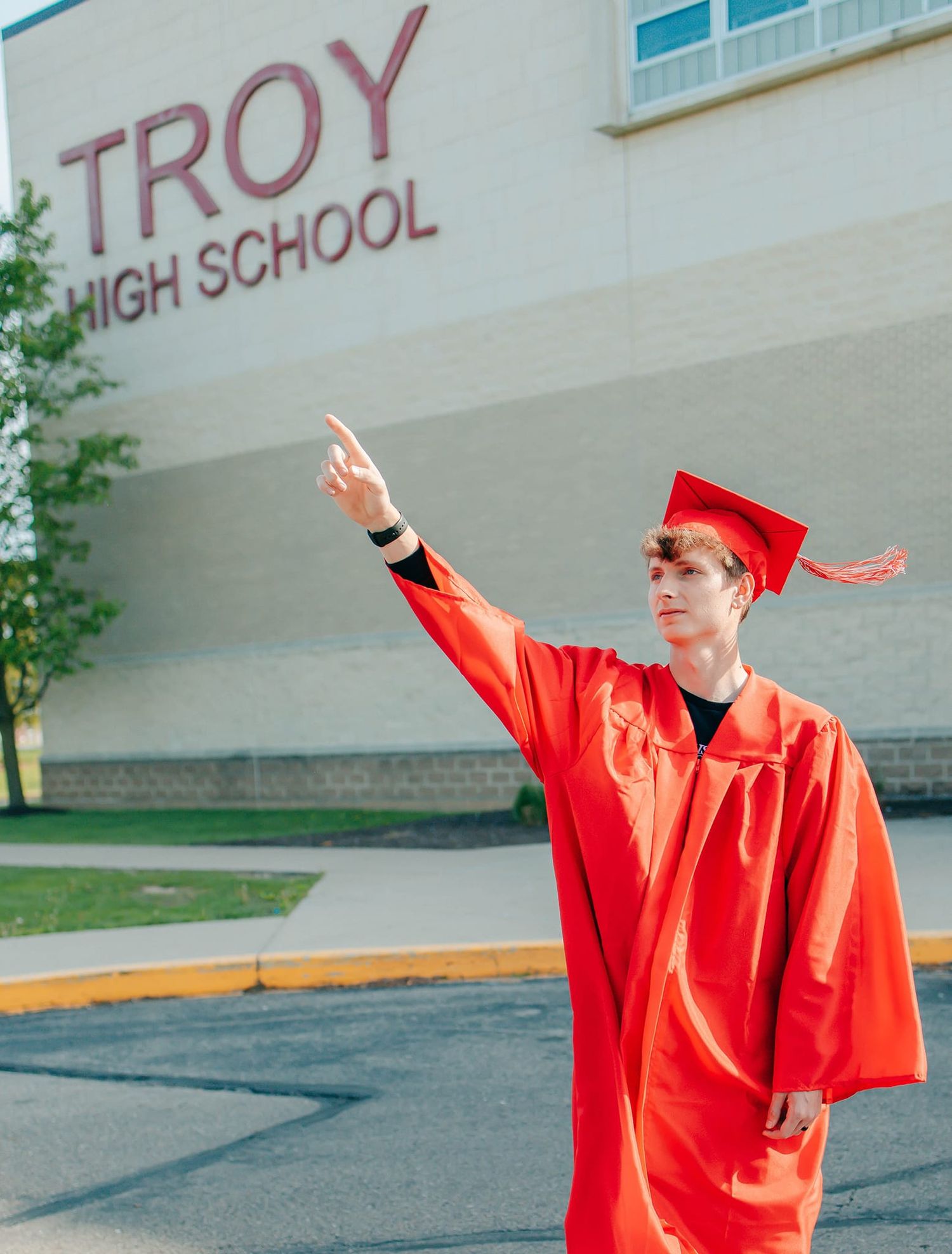 Graduate in bright red cap and gown poses triumphantly outside Troy High School.