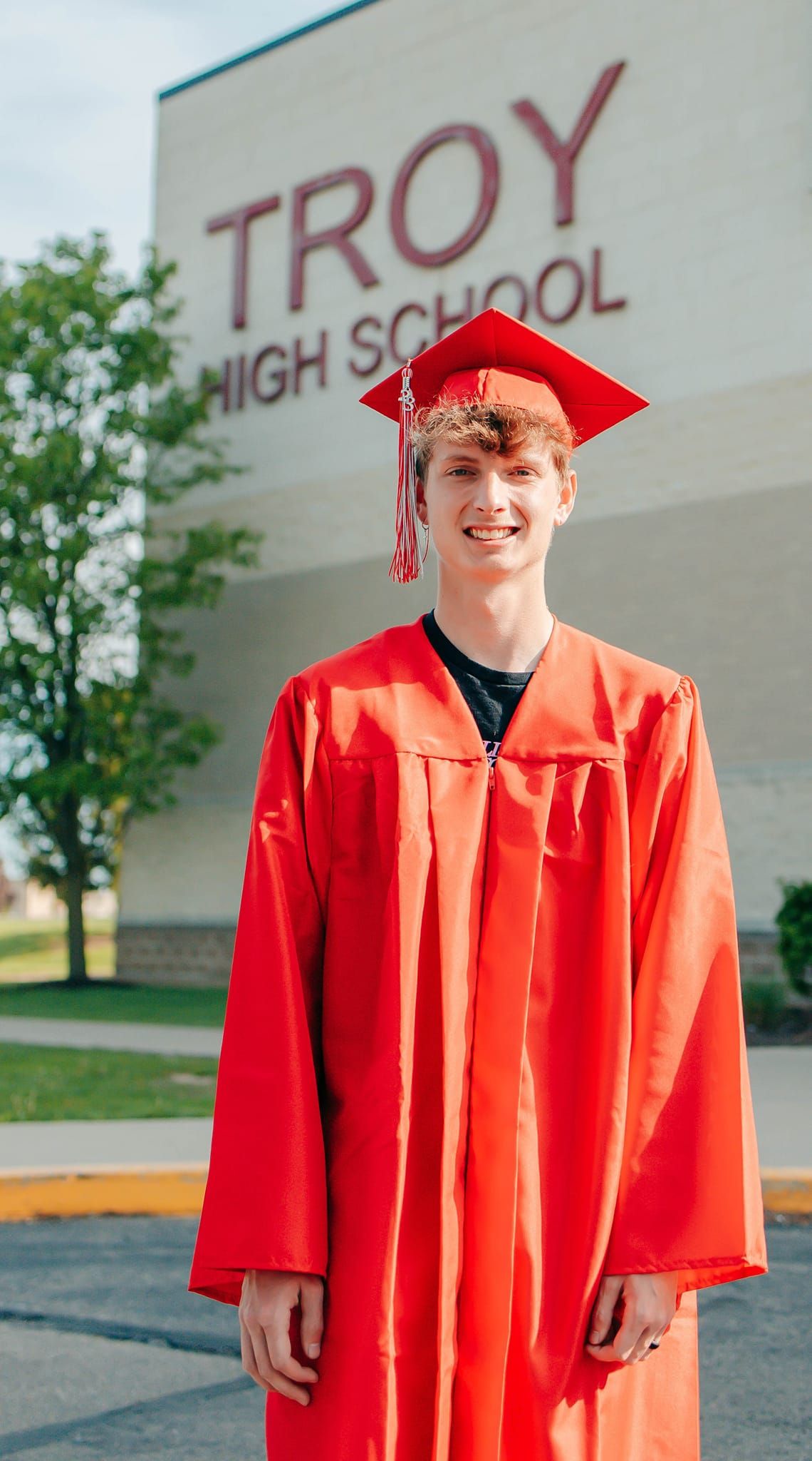 A graduate in bright red cap and gown poses in front of Troy High School.