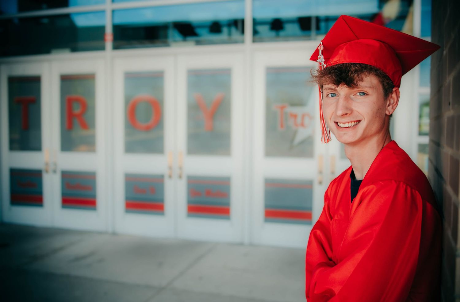 Graduate in red cap and gown smiles in front of building with red lettering.