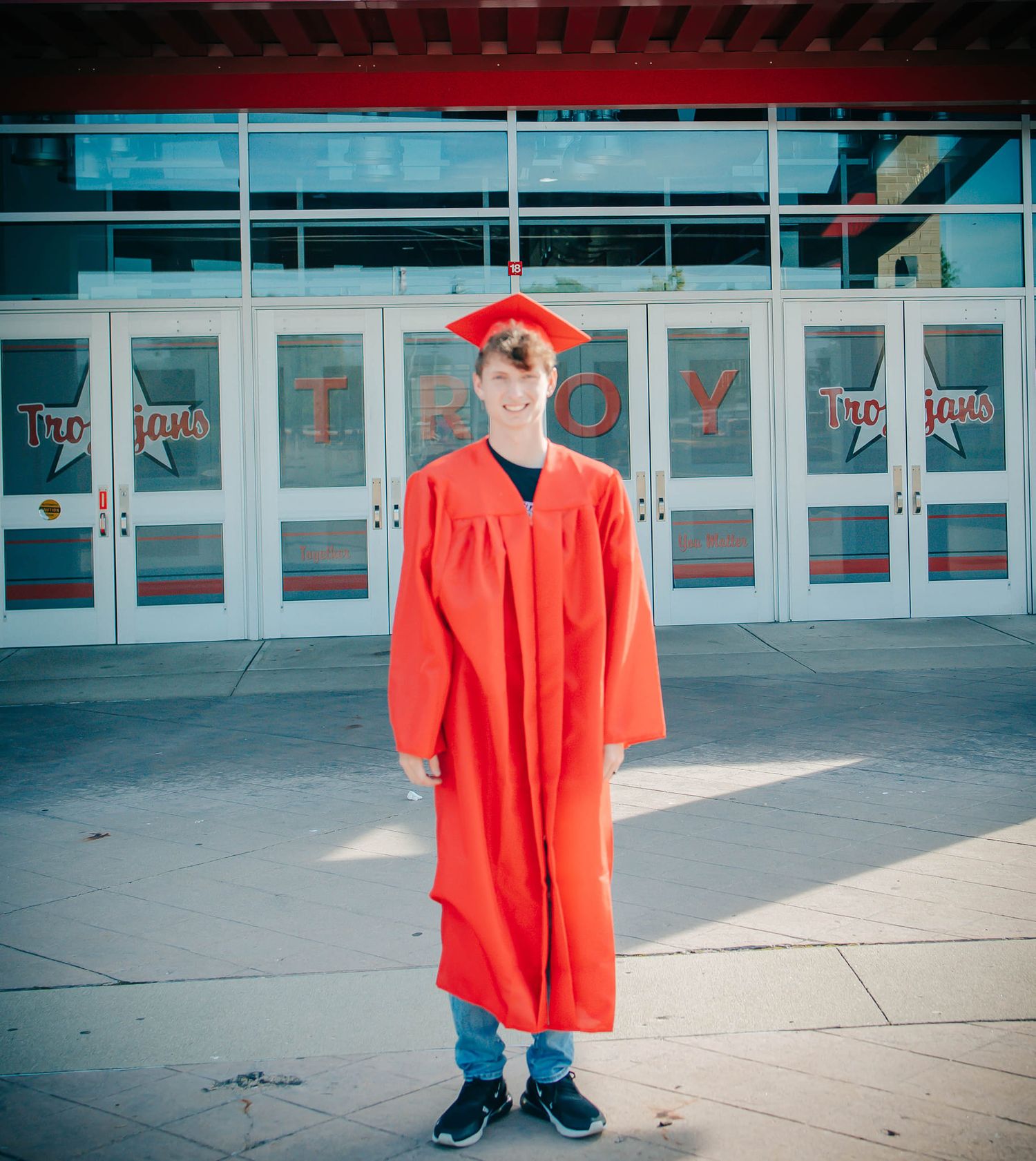 A graduate in red regalia stands in front of school entrance doors.