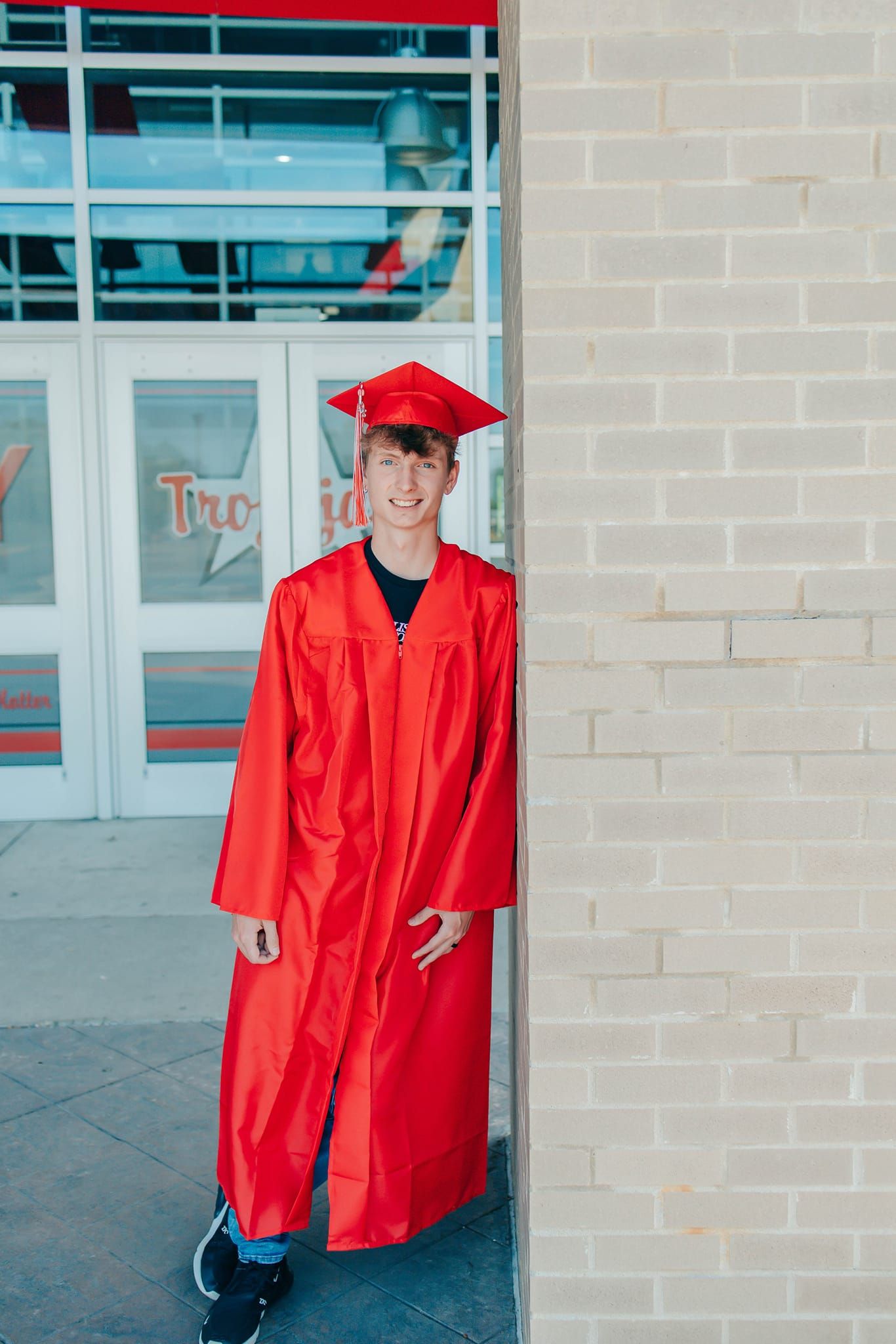Student in red graduation attire standing proudly outside school building entrance.