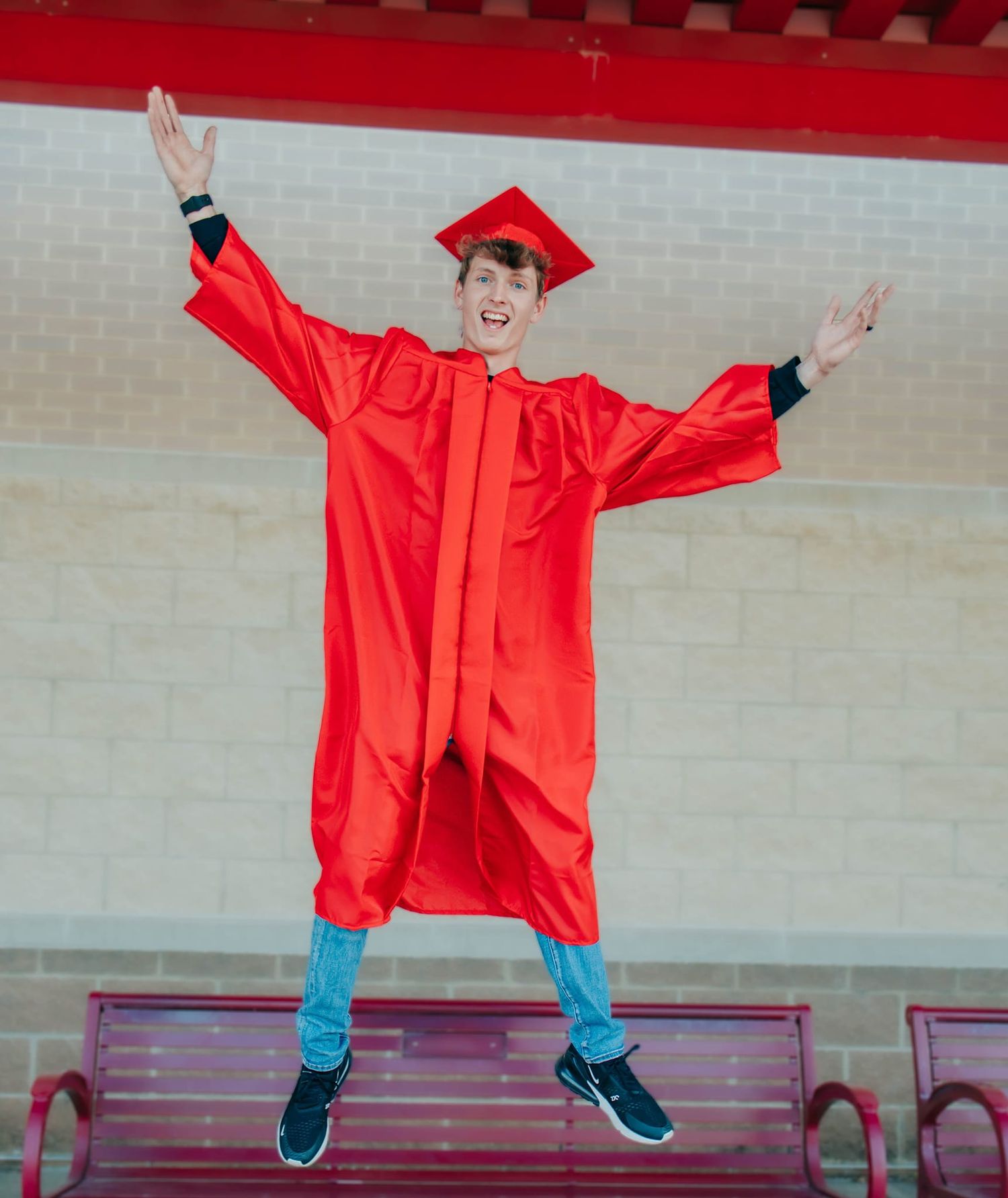 A graduate in red cap and gown strikes celebratory poses on a red bench.