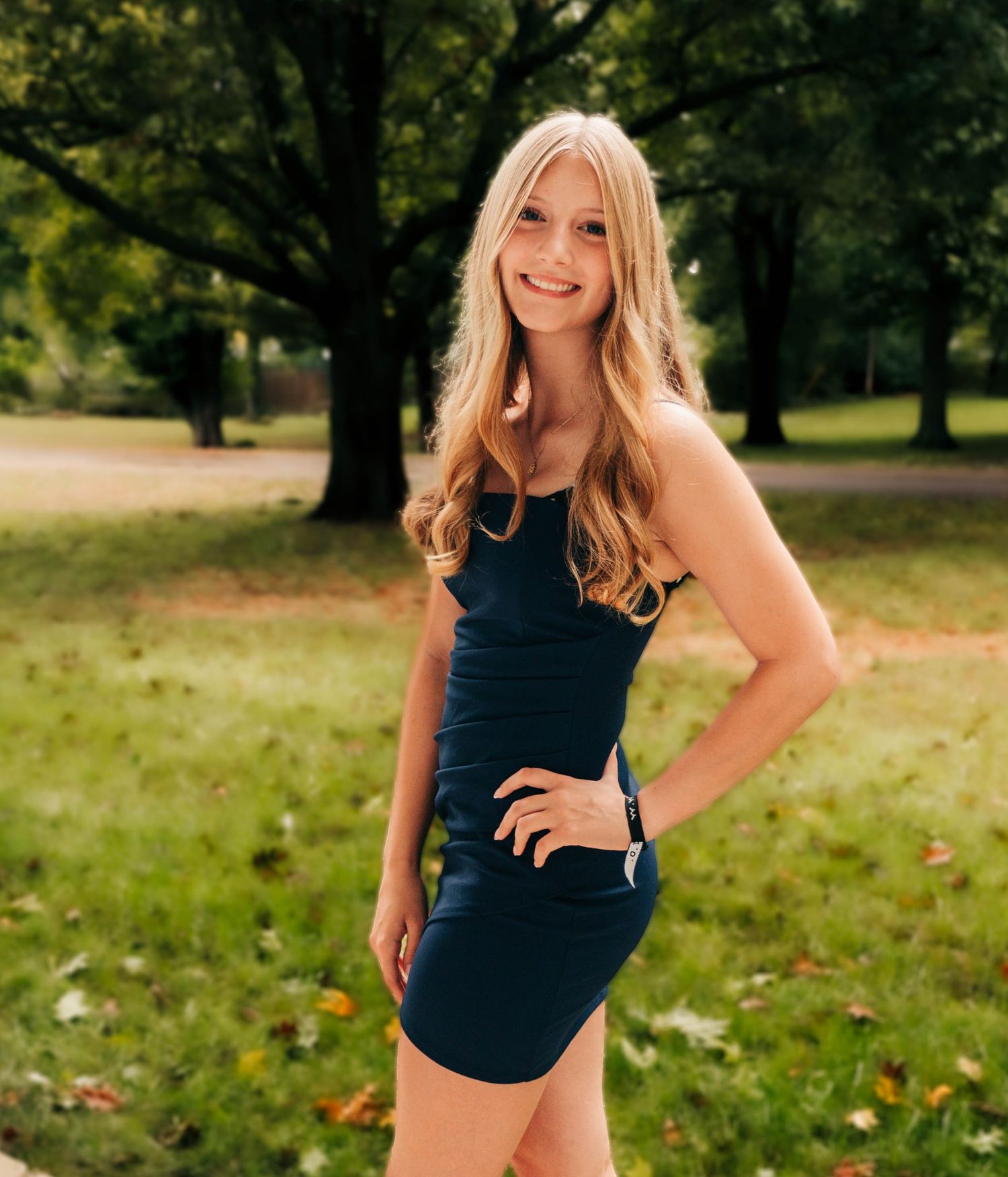 Person in navy sleeveless dress poses outdoors in park setting with autumn trees.