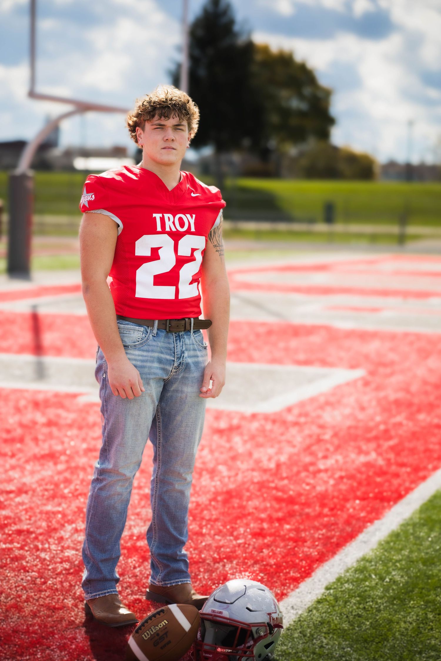 High school football player in red jersey and jeans poses with football on red and white field.