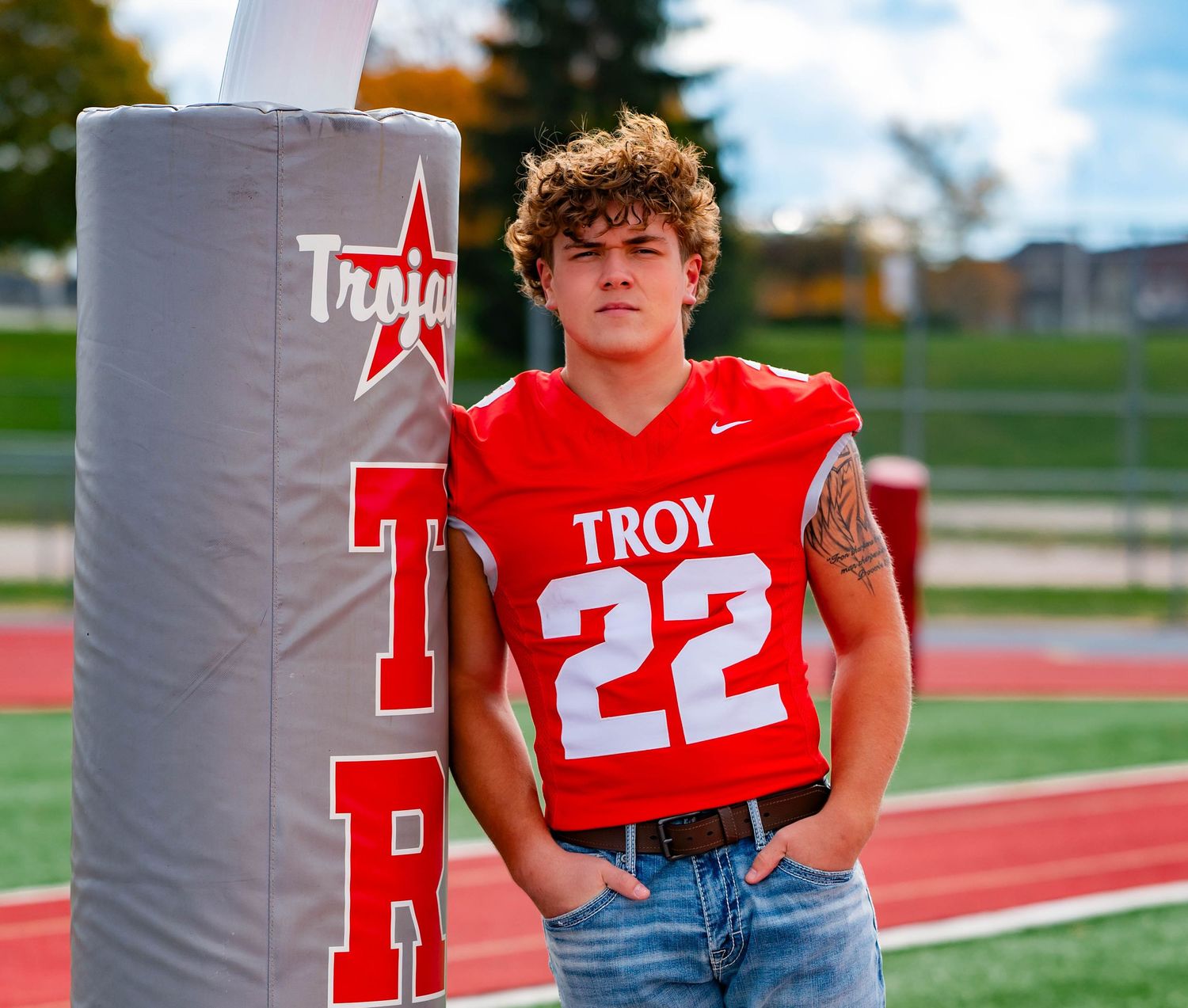 High school football player in red Troy jersey poses against padded goal post during outdoor photo session.