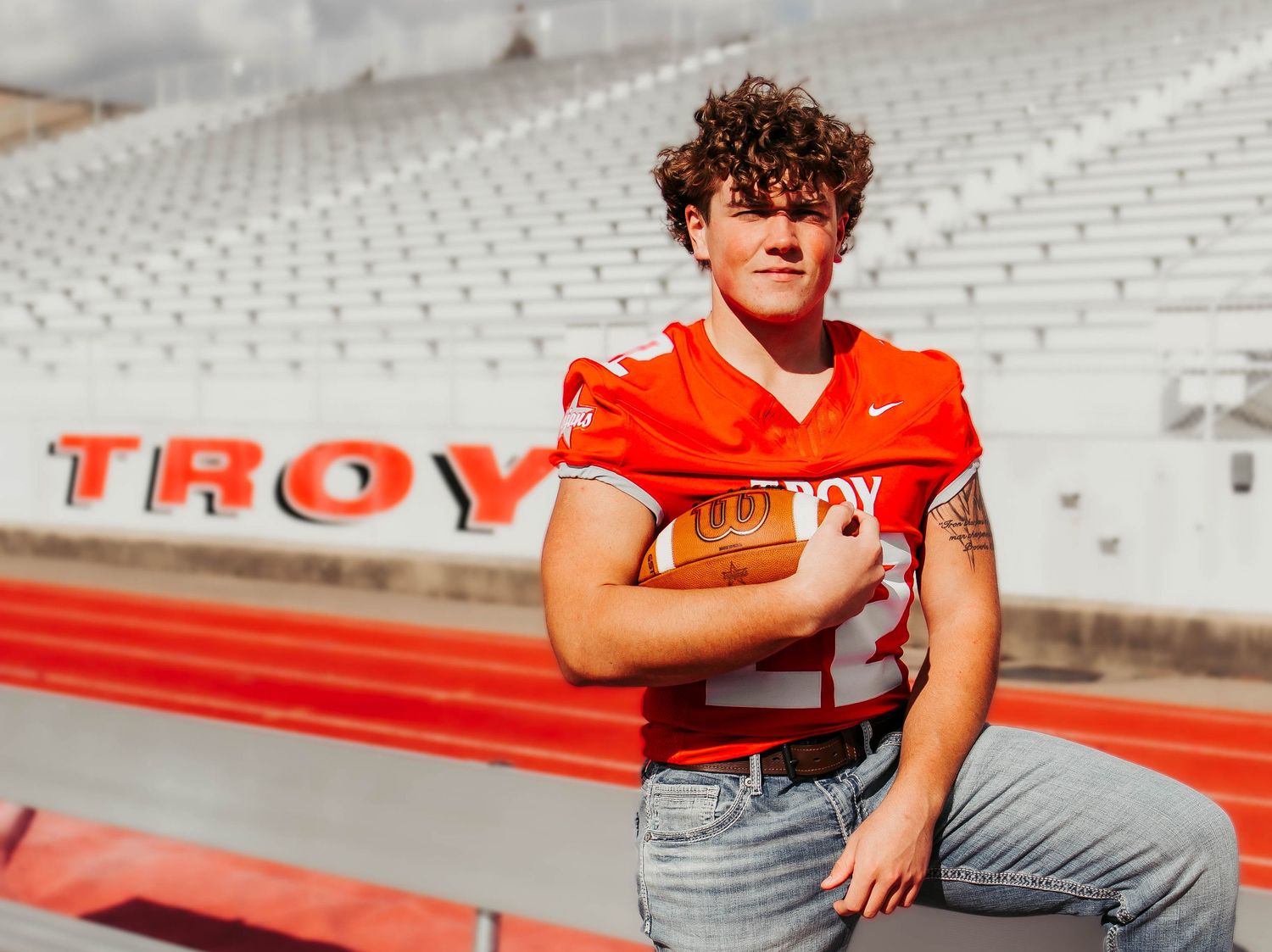 Football player in red jersey and jeans poses on stadium bleachers with TROY signage visible in background.