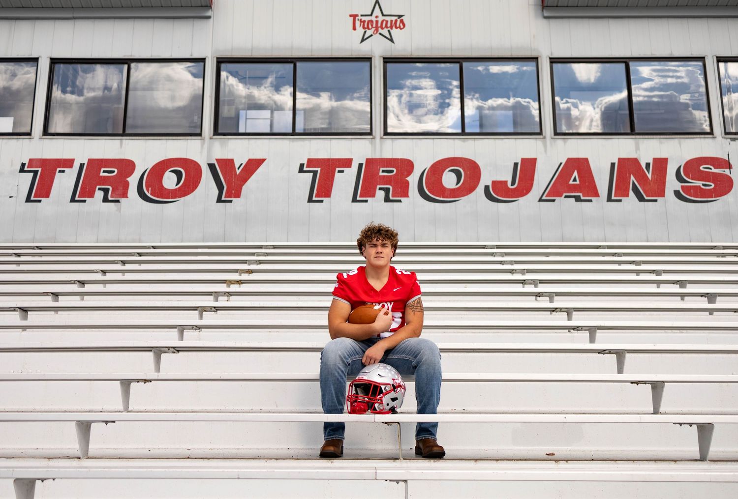 Student athlete in red jersey sits on bleachers beneath Troy Trojans signage at stadium.