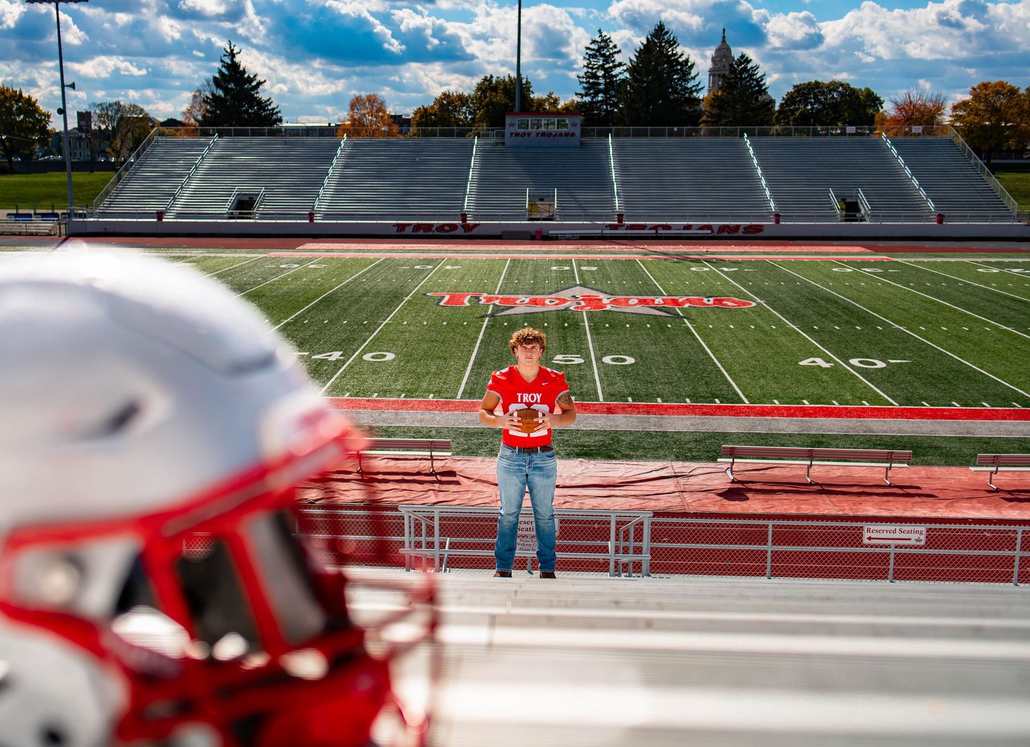A football helmet sits in the foreground overlooking an empty high school stadium field with red bleachers on a sunny day.