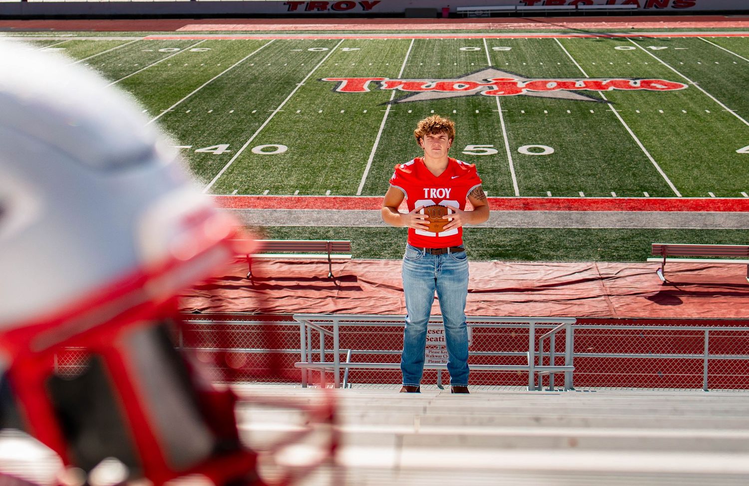 A panoramic view of empty red stadium bleachers and a football field with white yard lines and markings.