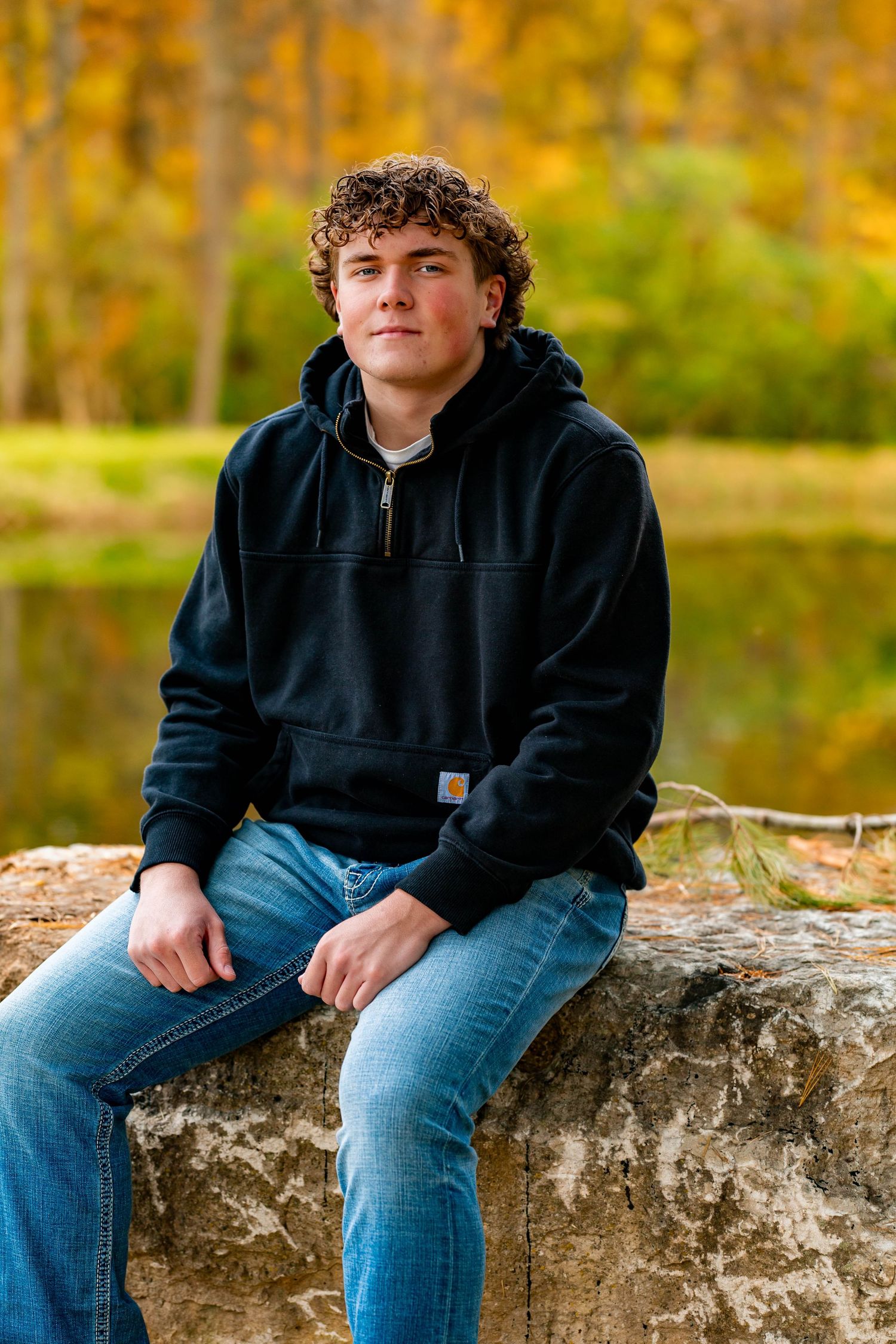 Person in black Carhartt hoodie and jeans sits casually on stone wall during autumn.
