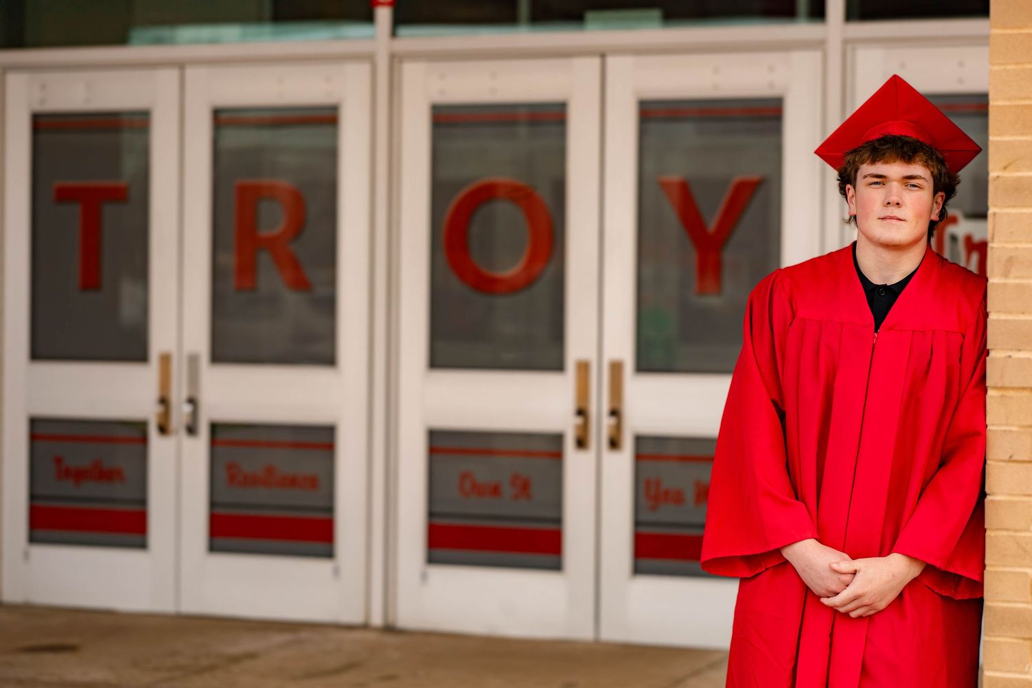 Graduate in bright red cap and gown stands in front of doors with TROY lettering at university entrance.