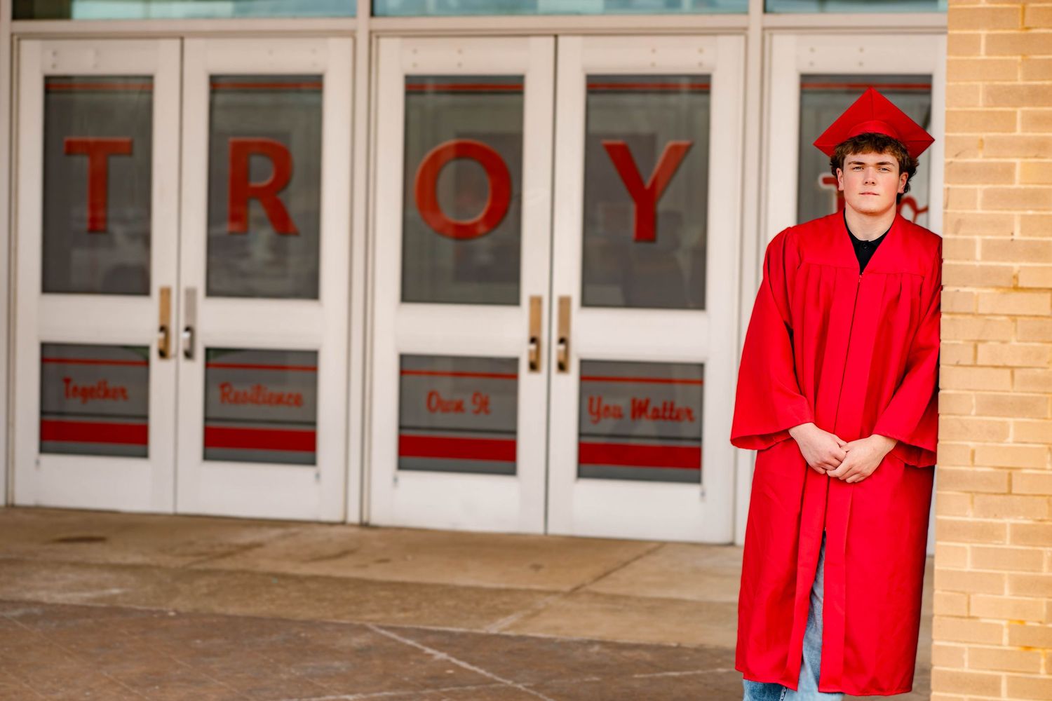 A person wearing a bright red graduation gown and cap stands in front of brick walls and glass doors.