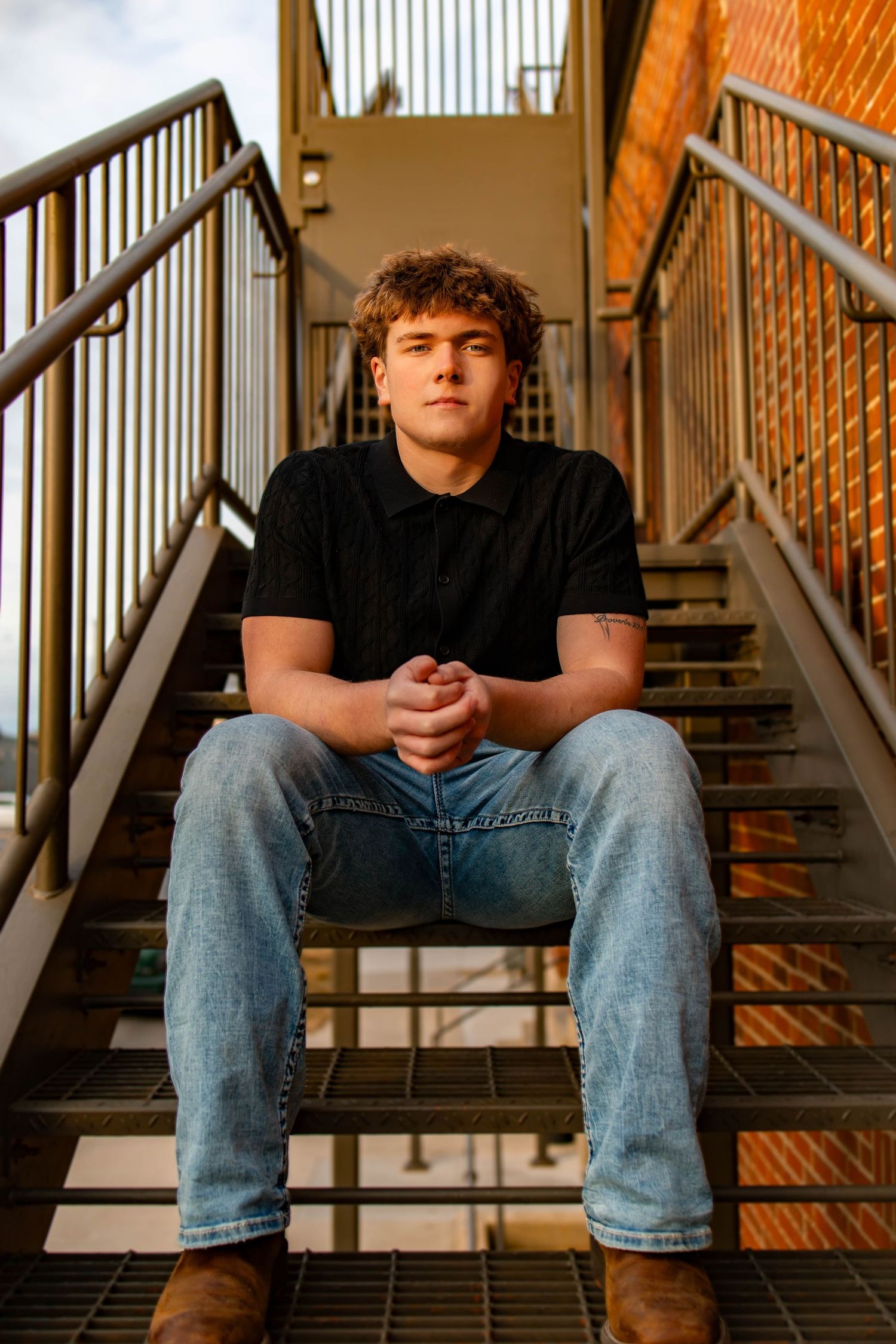 Person wearing black t-shirt and jeans sits thoughtfully on industrial metal stairs in warm lighting.