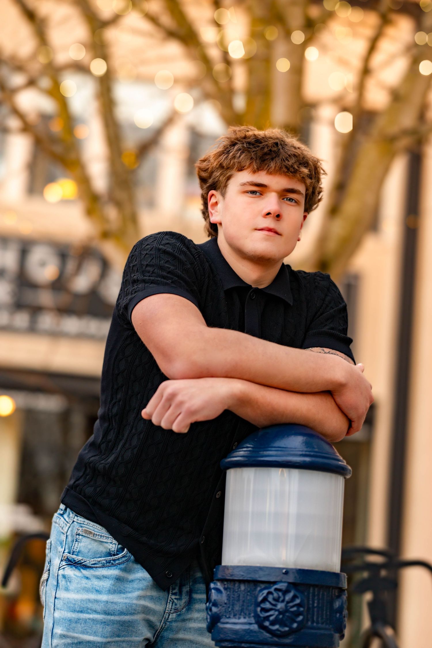 Student in black shirt and jeans leans on container in outdoor setting with bokeh lights.