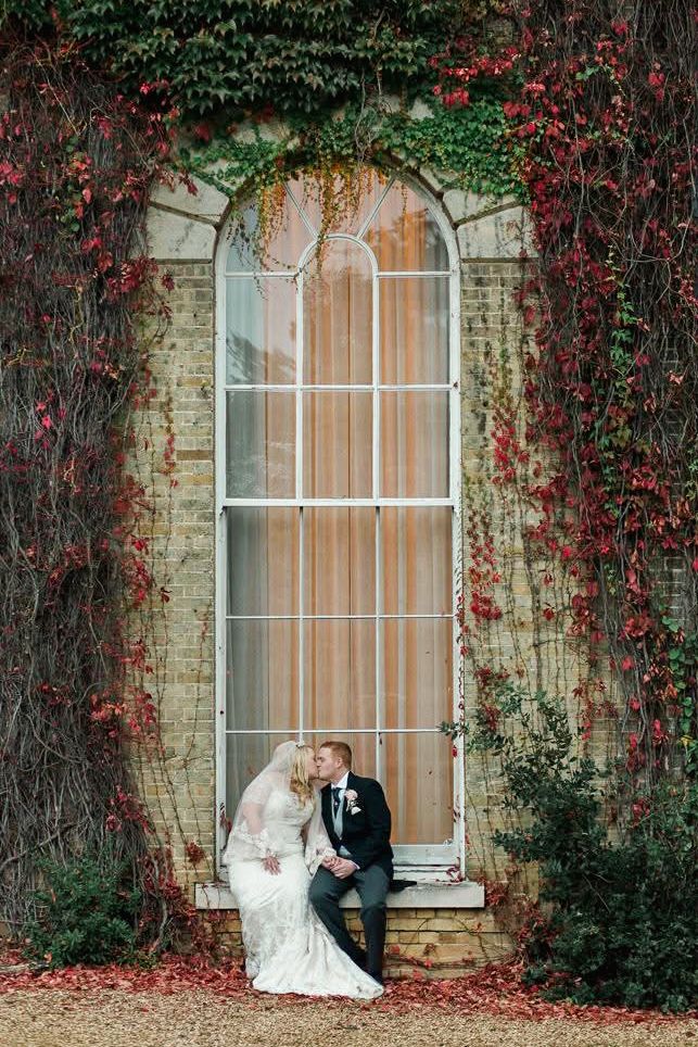 Romantic wedding couple poses against ivy-covered stone wall with red autumn foliage in elegant outdoor setting.