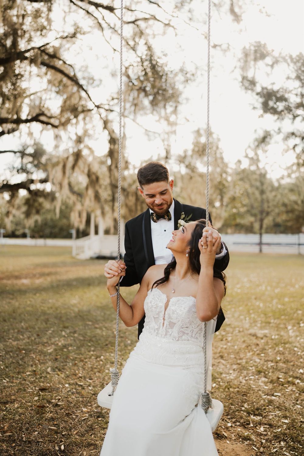 A couple in wedding attire share a romantic moment on a tree swing in a rustic outdoor setting.