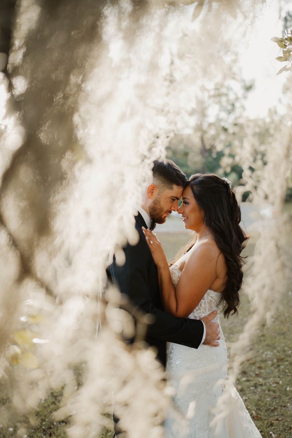 A romantic wedding couple embraces in a dreamy outdoor setting with soft sunlight filtering through trees.