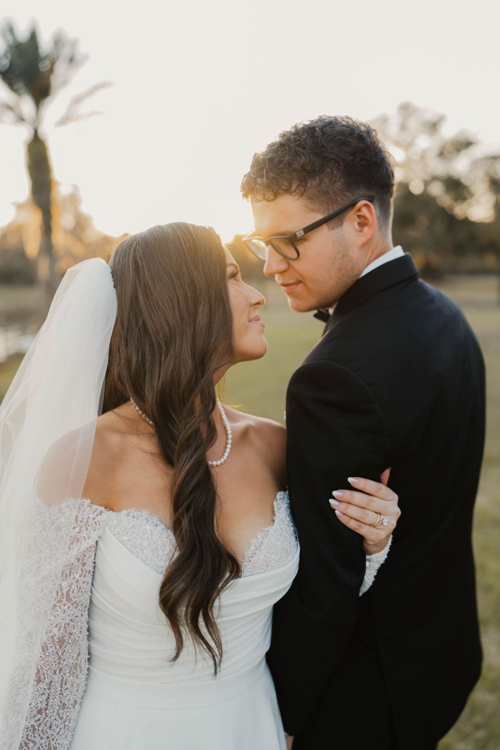 A romantic sunset wedding portrait of a couple embracing outdoors, with the bride in a lace gown and long veil.