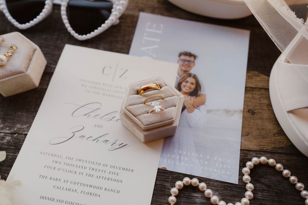 Wedding invitation suite with pearls, rings, and delicate stationery details arranged on rustic wooden surface.