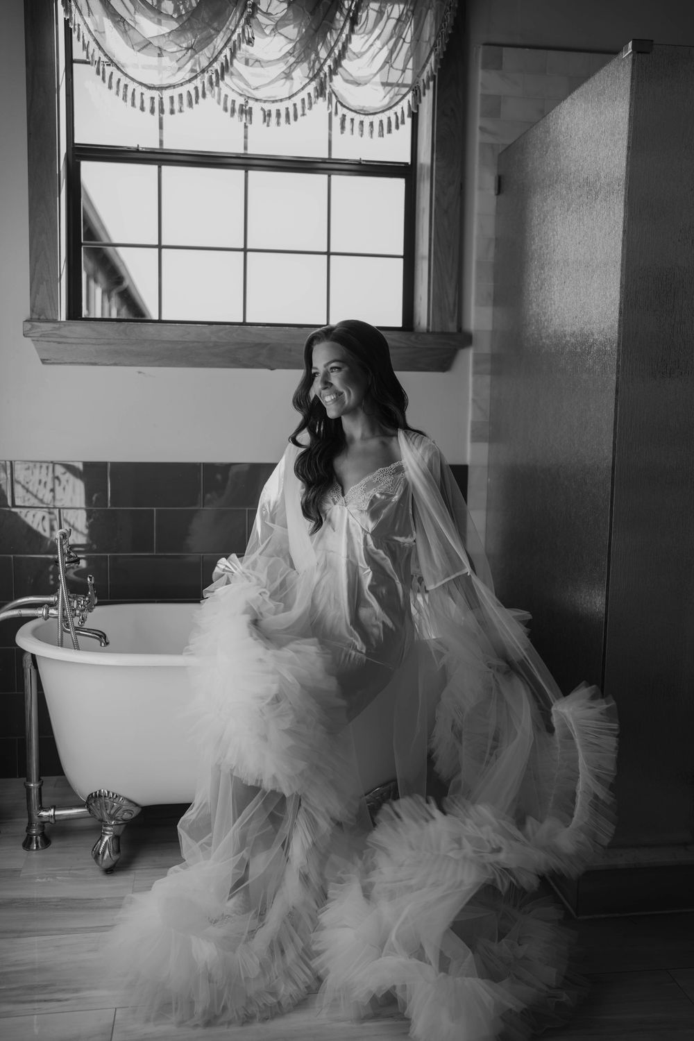 Black and white boudoir photo of a bride in a flowing robe sitting elegantly by a clawfoot bathtub near a window.