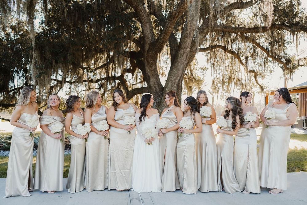 Bridesmaids wearing champagne colored dresses stand in line under Spanish moss draped trees.