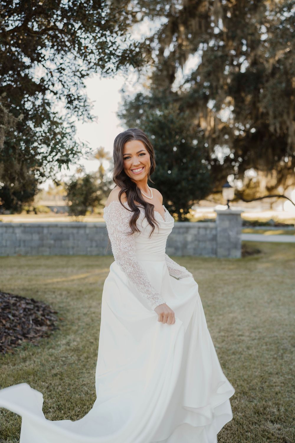 A bride in a flowing white lace wedding dress twirls joyfully outdoors under Spanish moss trees on a sunny day.