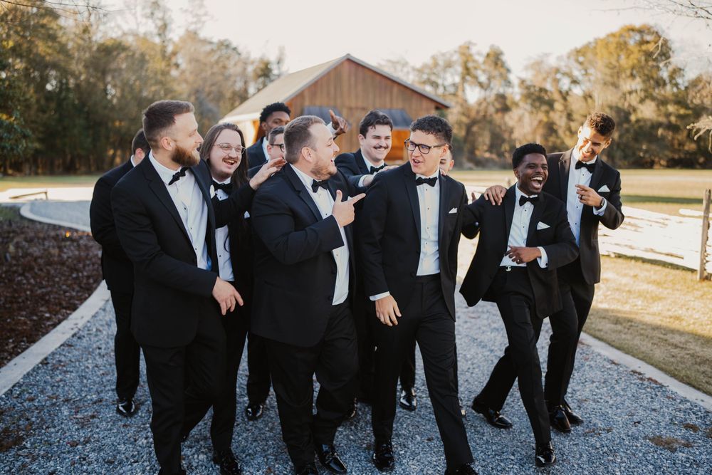 A group of groomsmen in black tuxedos laugh together on a gravel path with a rustic barn in the background.