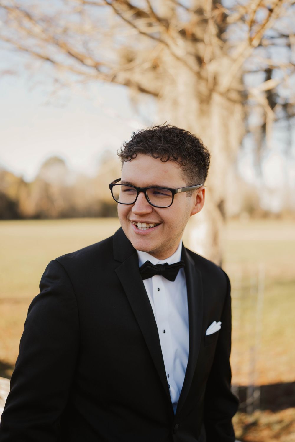 A person in a black tuxedo and bow tie smiles warmly while wearing glasses during an outdoor formal photoshoot.