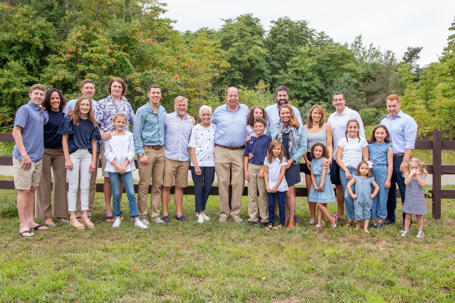 Large multigenerational family group portrait taken outdoors on a green lawn near a wooden fence.