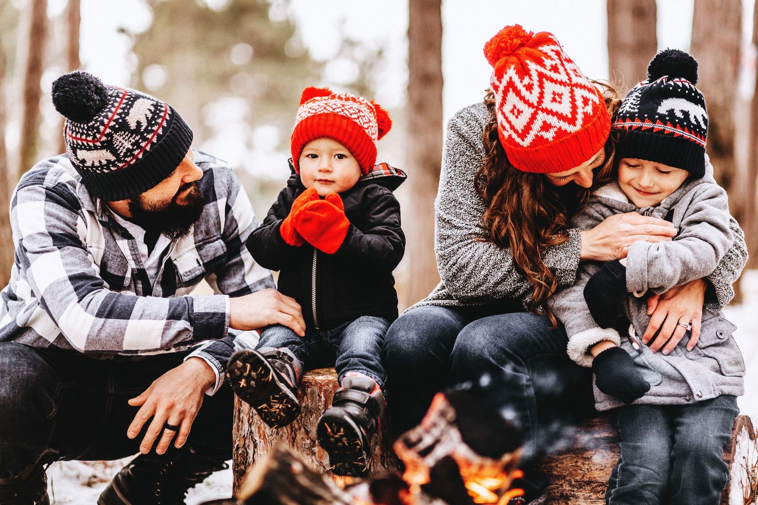 Husband, wife, and their two boys cuddle near a fire at the tree farm mini session. They are wearing winter hats with poms on top