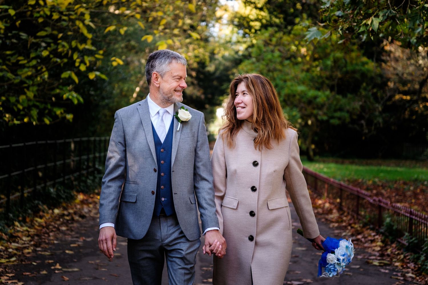 Couple walking together in autumn park wearing formal attire and holding hands.