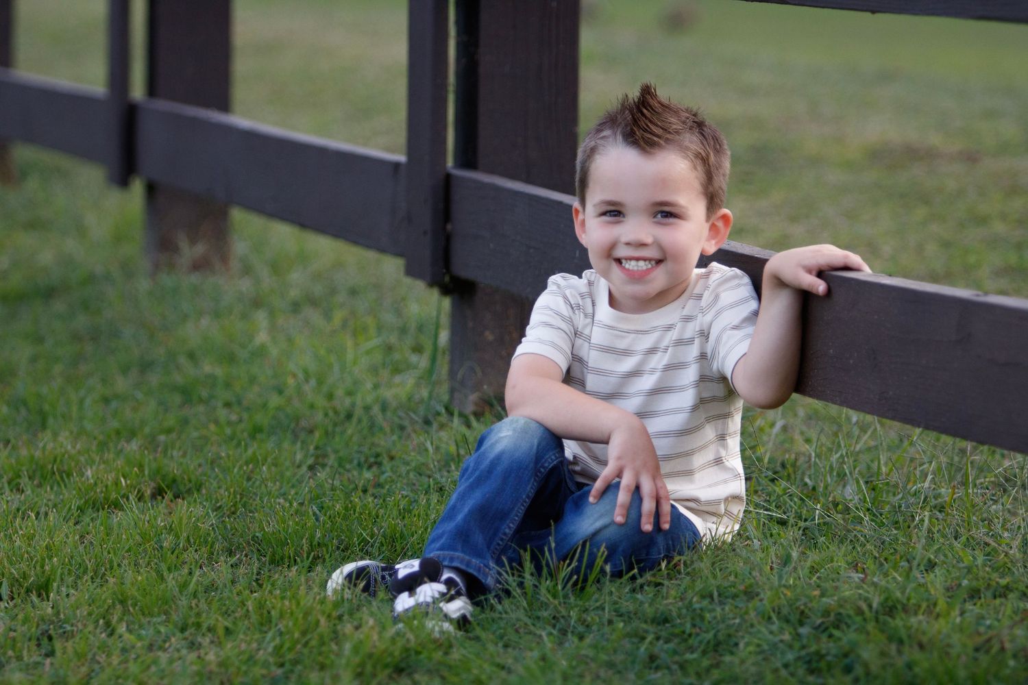 A playful child lies on green grass making silly expressions while wearing a striped shirt and jeans.