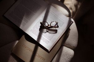 Reading glasses rest on an open book page with dramatic shadows and lighting creating a moody literary scene.