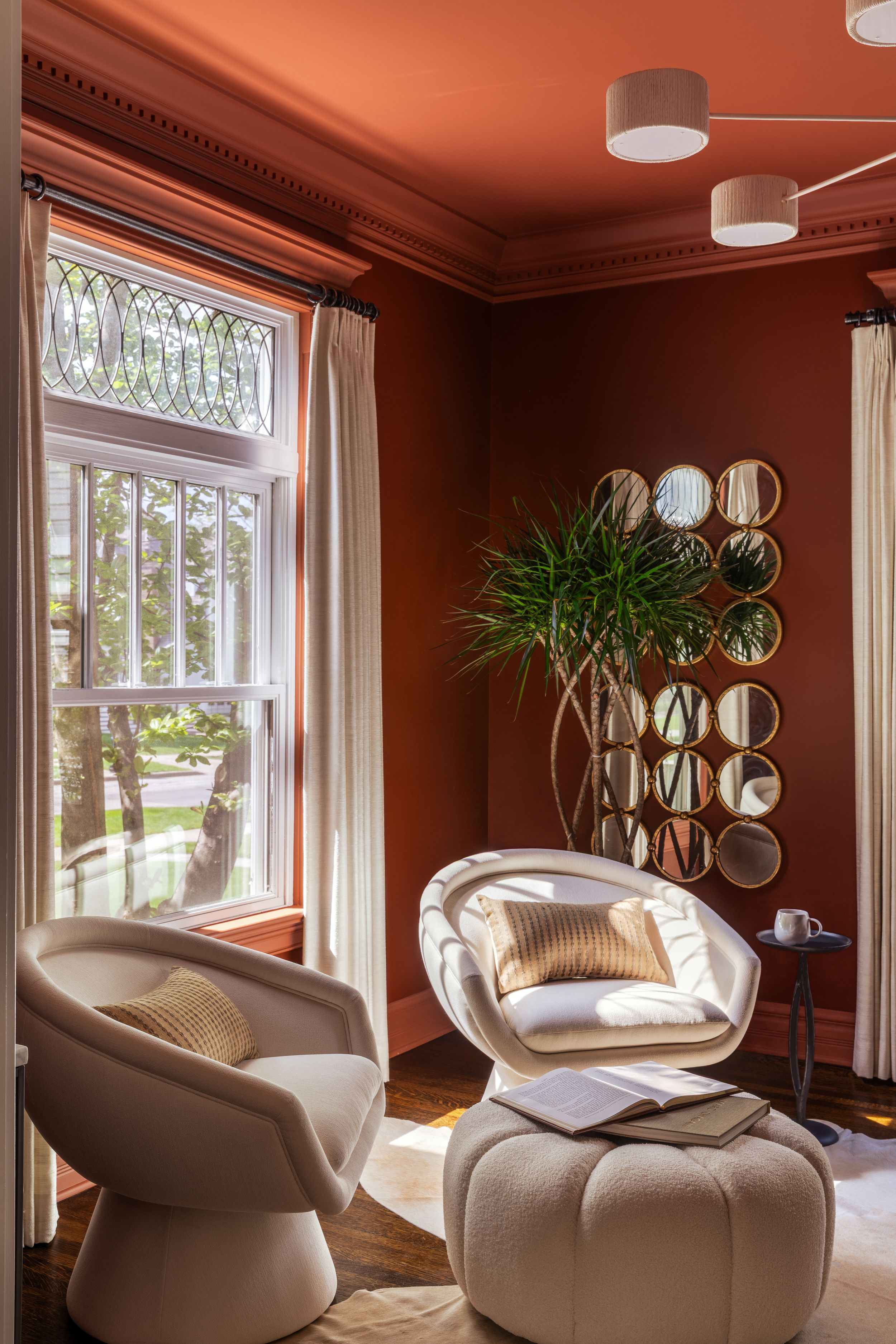 Modern living room corner with cream swivel chairs, round ottoman, decorative mirrors, and large window with ornate transom.