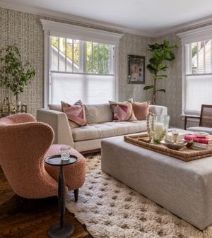 Cozy living room with beige sofa, coral accent chair, textured wallpaper, large windows and indoor plants.