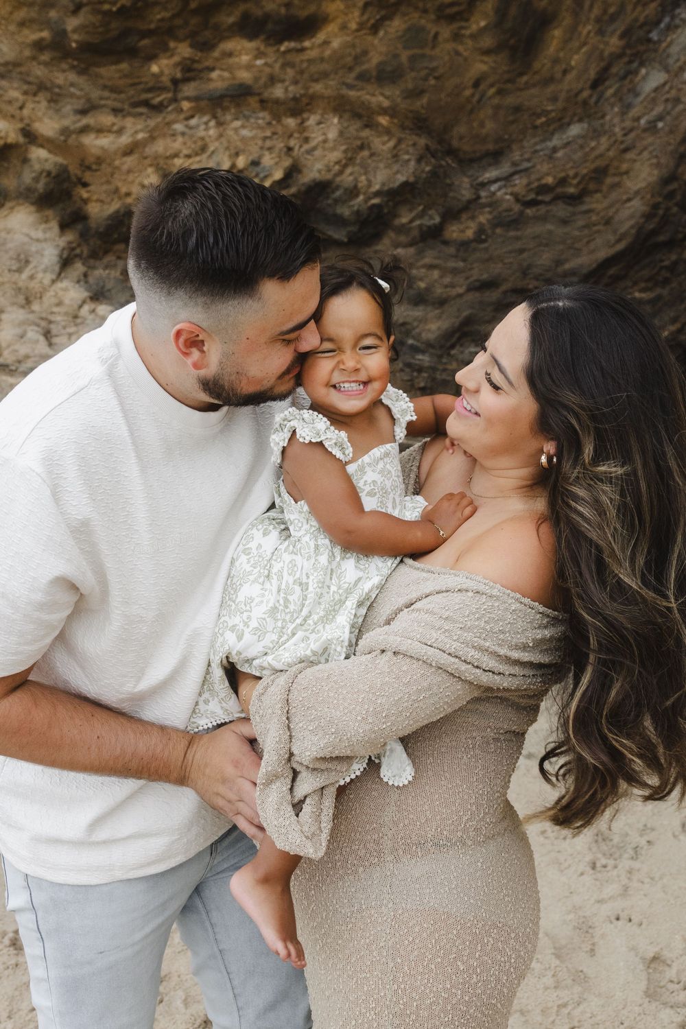 A family shares a tender moment together against a rocky backdrop at the beach, wearing neutral colored clothing.
