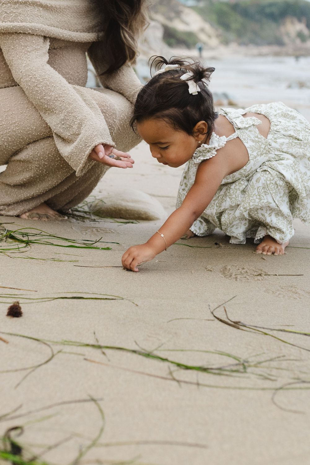 A small hand draws in the sand on a beach while wearing a flowy dress.
