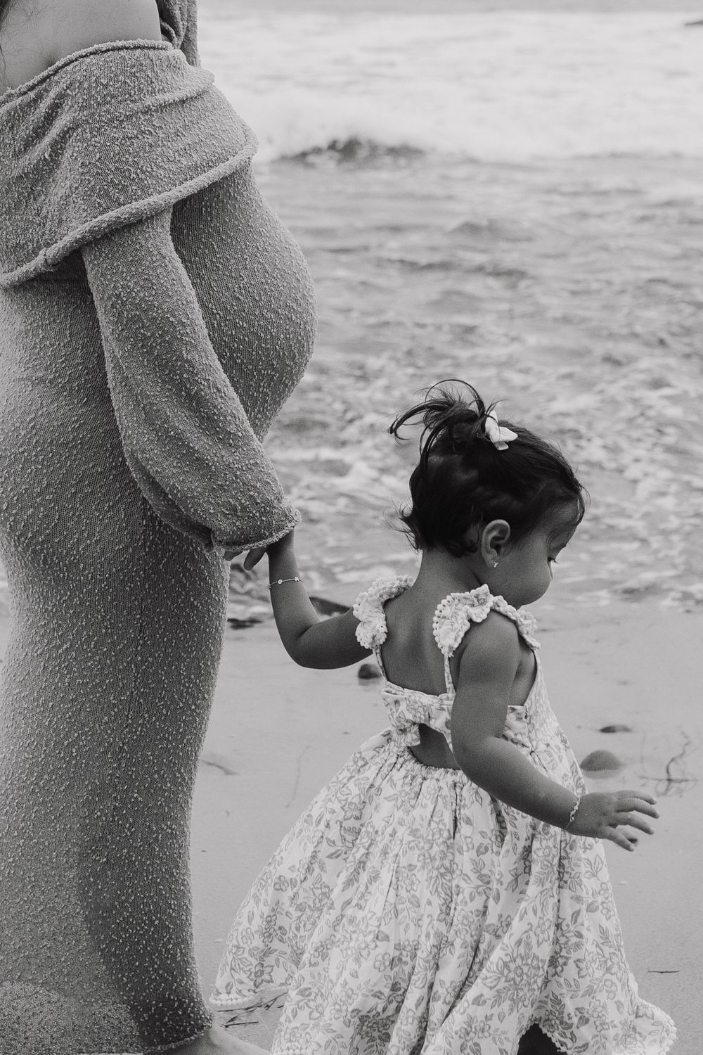A child in a floral dress walks along the beach while holding hands with someone in a cozy sweater.
