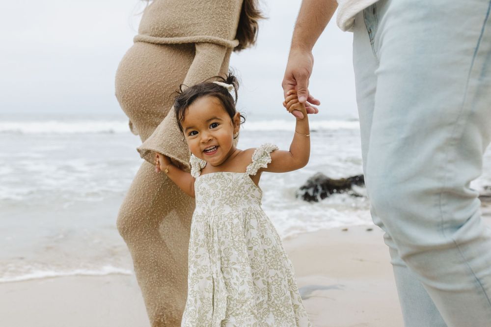 A family holds hands while walking along the shoreline in coordinated neutral outfits.