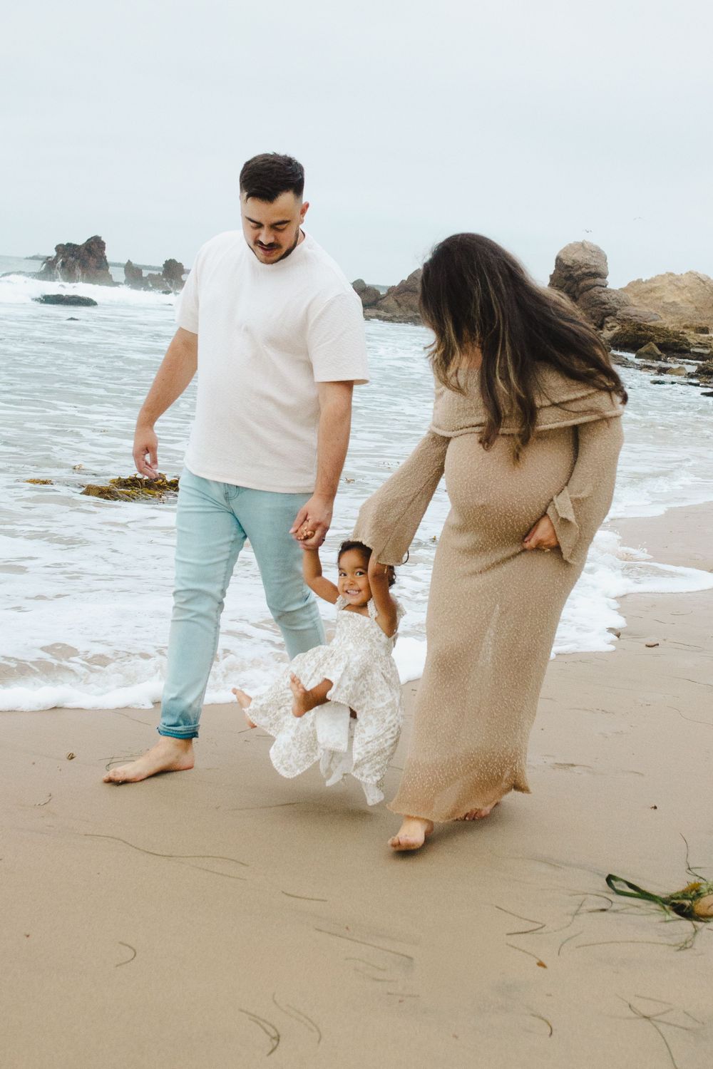 A family plays together on a rocky beach, lifting a small child while waves crash in the background.