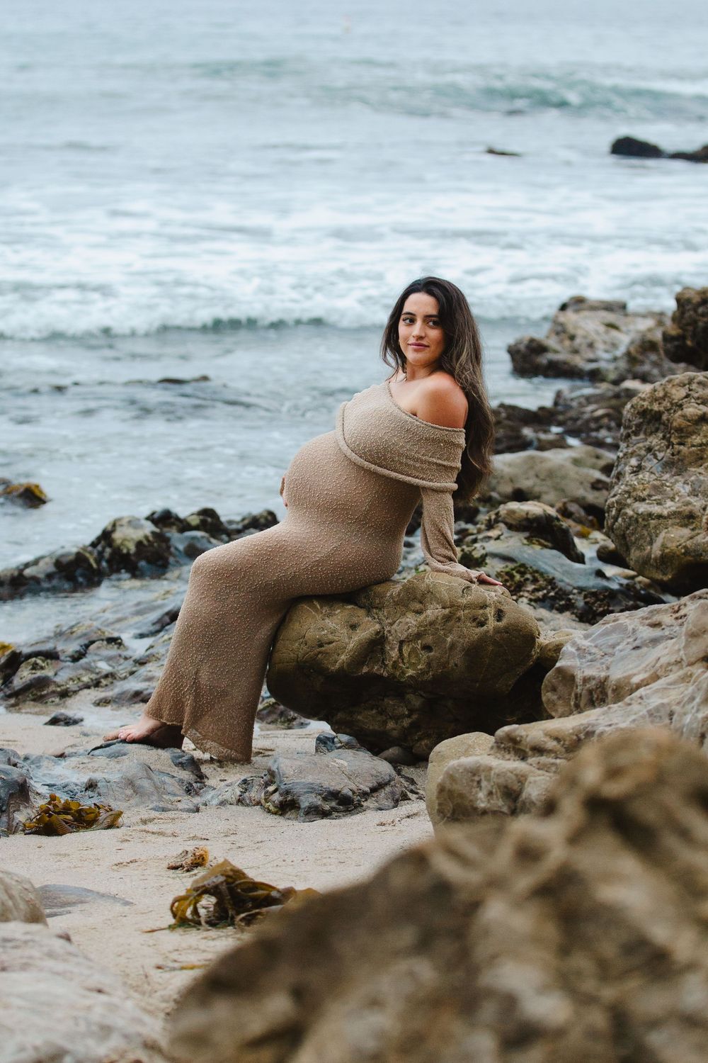 Woman in beige knit dress poses on rocky beach with ocean waves in background.