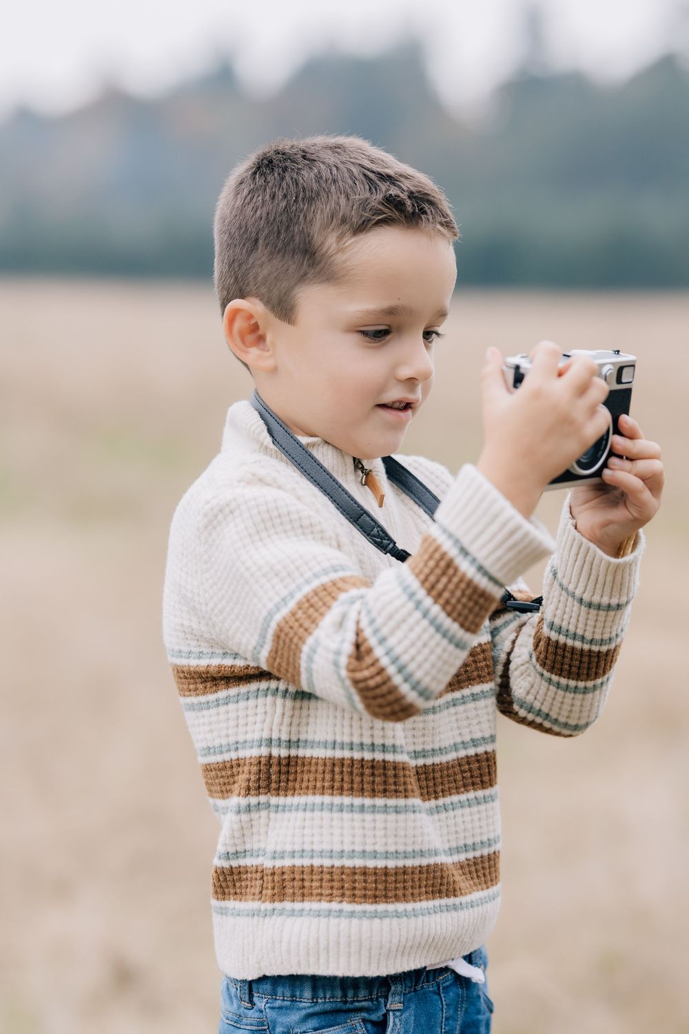 Young boy in striped sweater holding a vintage camera in an autumn field.