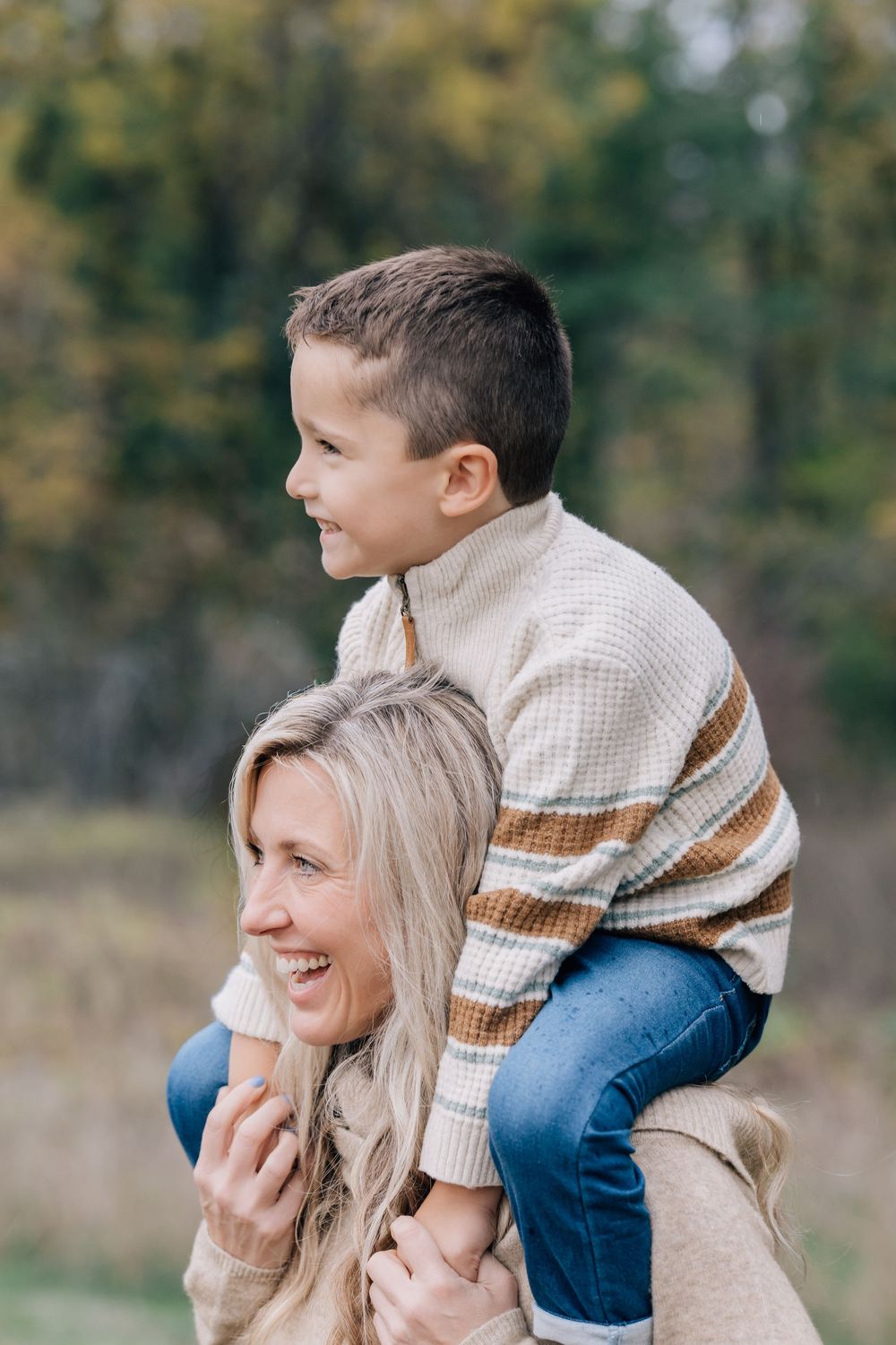 Blonde mother laughing with young son sitting on her shoulders in autumn foliage.