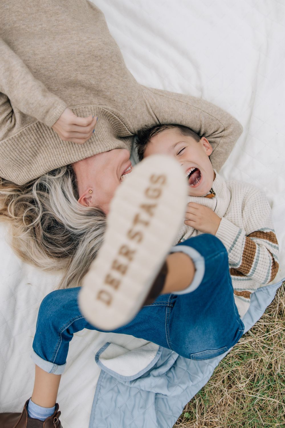Mom and child laughing together on white blanket outdoors, child wearing Deer Stags shoes.