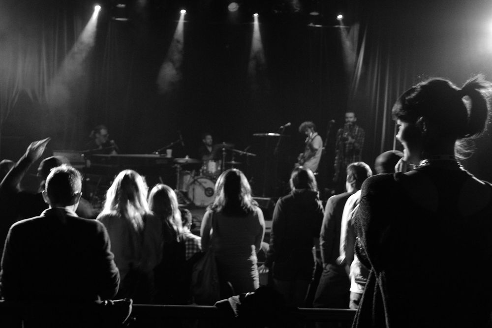 Black and white photo of a crowd at a concert venue with stage lighting silhouetting the audience members.