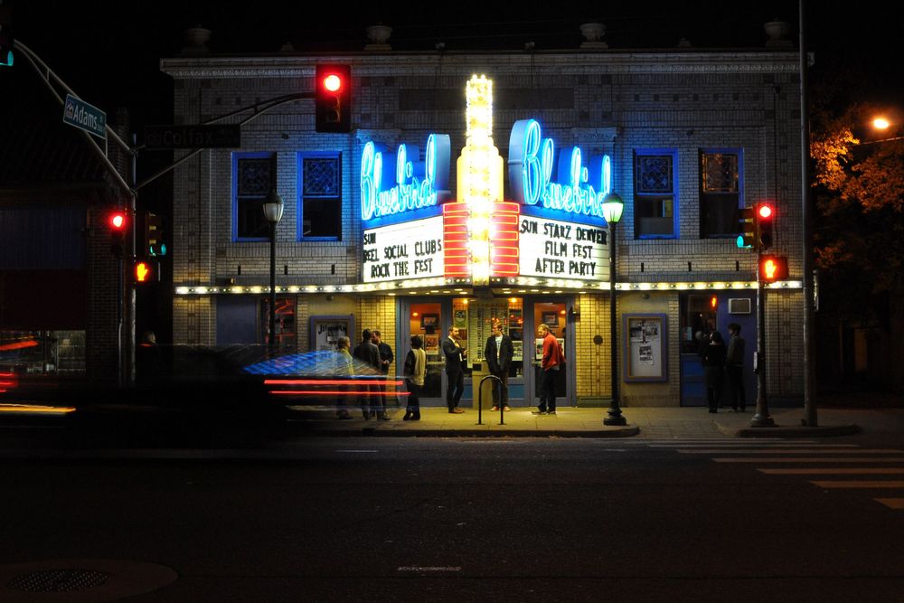 Illuminated vintage movie theater facade with neon blue crown signs glows at night on a city street corner.