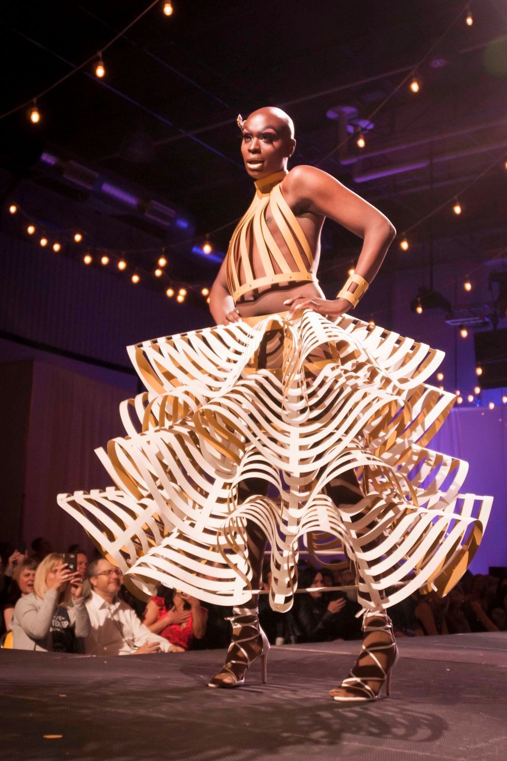 A dancer performs in a white ruffled costume creating a dynamic spiral motion effect on a dark stage with string lights above.
