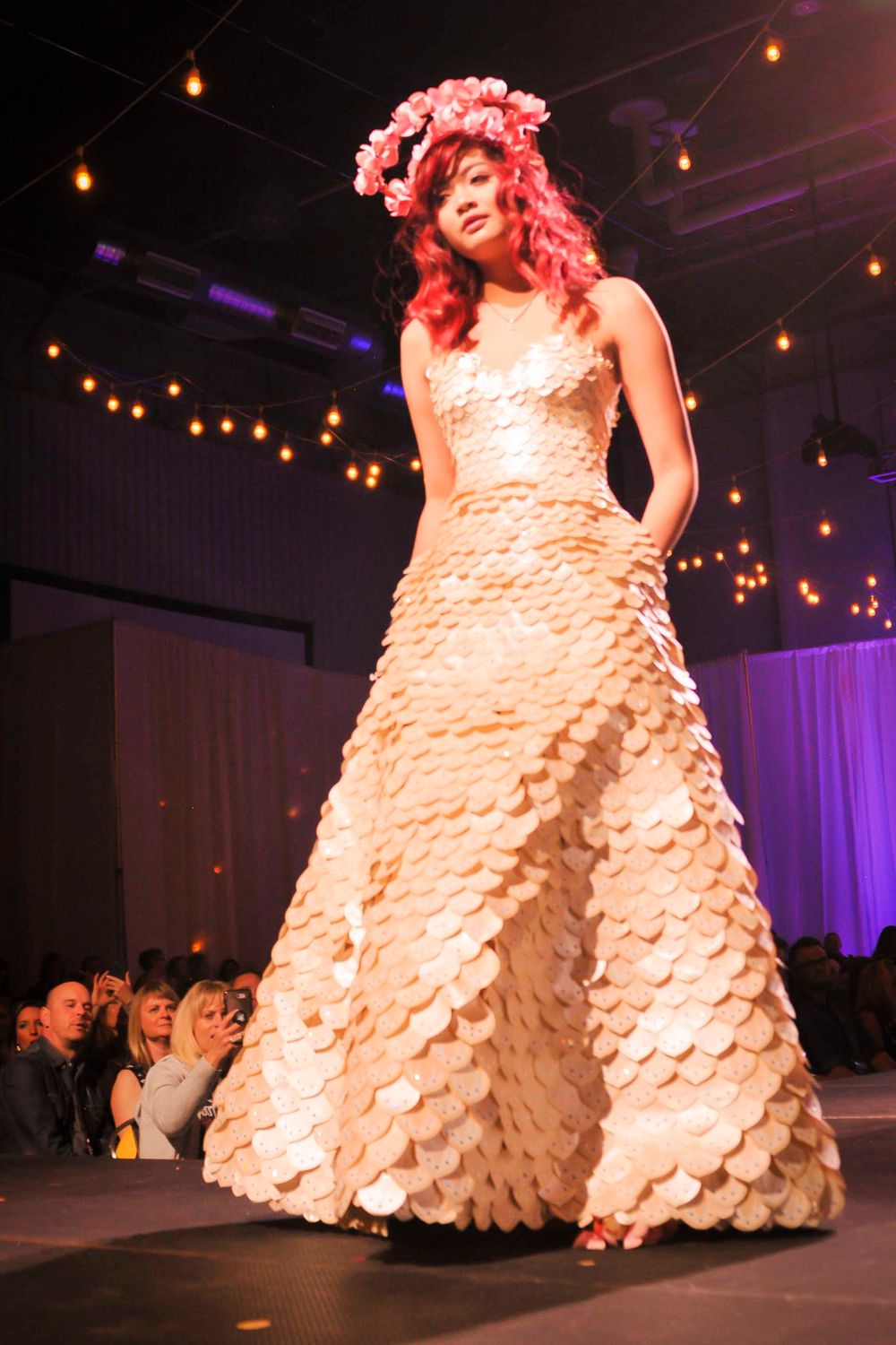 A model walks the runway in a dramatic white ruffled ballgown under warm lighting at a fashion show.
