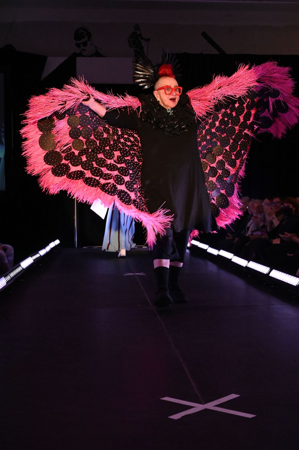 Pink feathered costume wings with black honeycomb pattern displayed on dark runway during fashion show.
