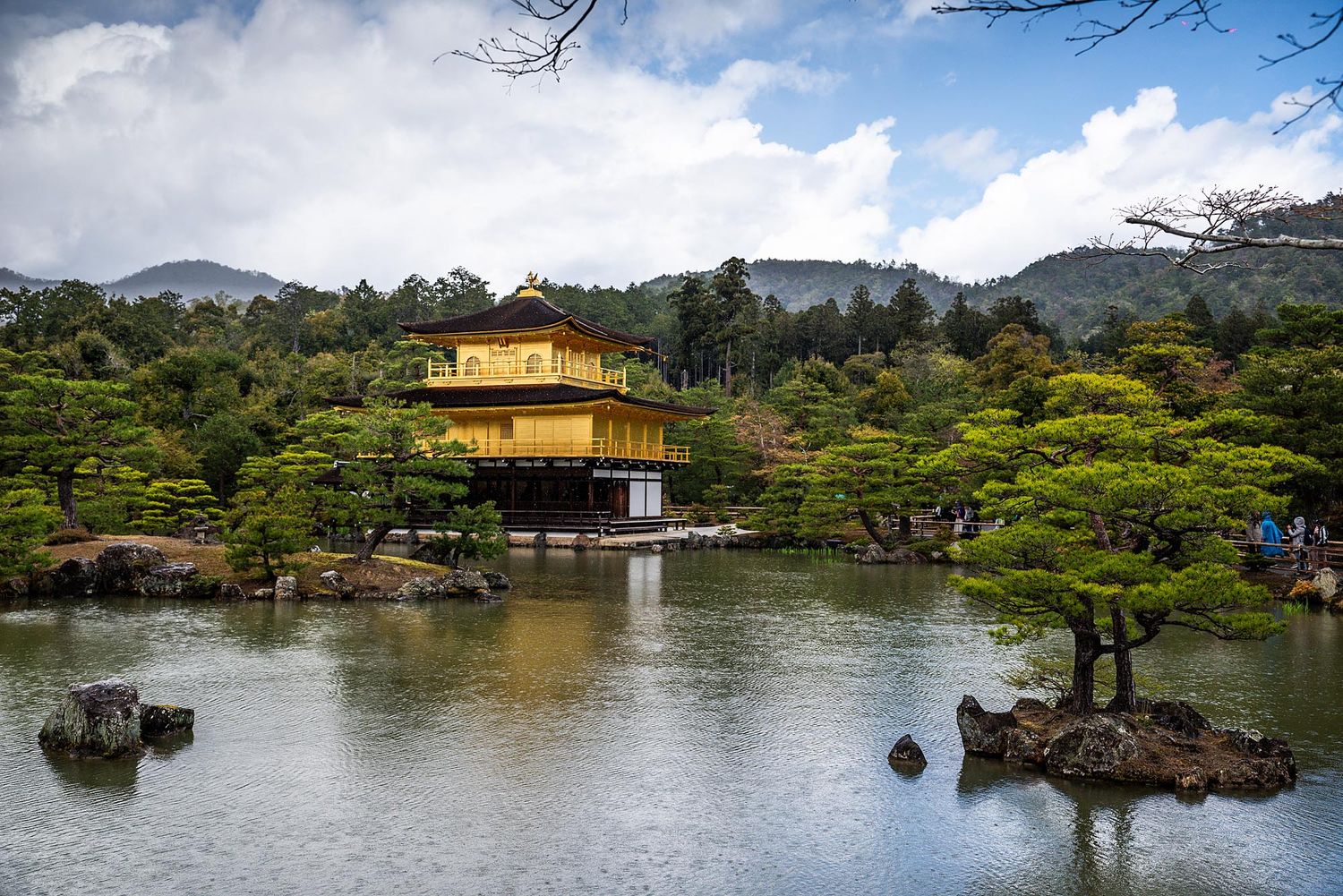 A golden pavilion temple reflects in a tranquil lake surrounded by Japanese gardens and mountains.